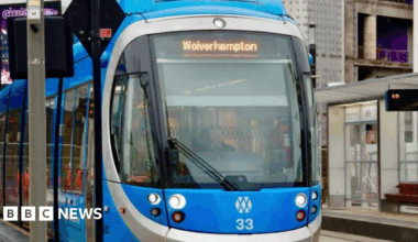 A tram navigates it way through Birmingham city centre amid major construction work with cones also laid out on roads. A person walks along a footpath to the left of the image.