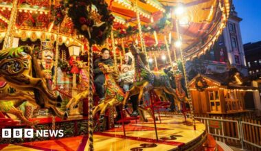 A lit up funfair roundabout as evening falls with people sitting astride tow of the ride's horses