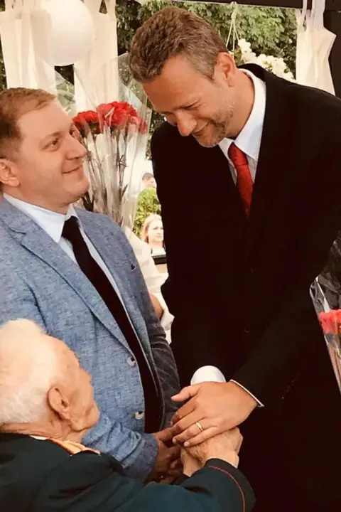 Telegram Two man are standing up and are greeting another older man who is sat down. The white-haired man who is sat down is wearing a black suit and is shaking hands with both the men that are standing. The brown-haired man on the left is wearing a light blue suit, white shirt and dark tie while the brown-haired man on the right has stubble and wearing a dark suit, white shirt and red tie 