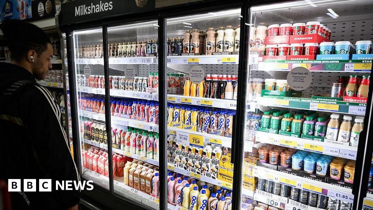A man with dark hair stands in front of a large supermarket fridge containing shelves of milk-based drinks, such as flavoured milkshakes and milky coffees like lattes, cappucinos and iced coffees.