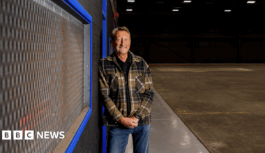 Stephen Knight is wearing blue jeans and a brown checked jacket. He is leaning against a wall