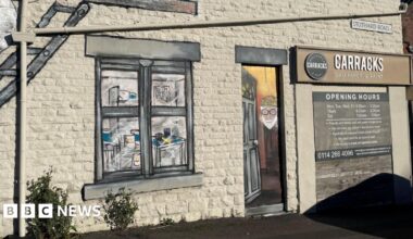 The exterior of bricked building. There is a large mural of a window  painted in a realistic style. To the right of the window mural is a painted door featuring a portrait of an older person wearing glasses and a hat. Above the door is a black and white sign that reads 'Carracks Wallpaper & Paint'.