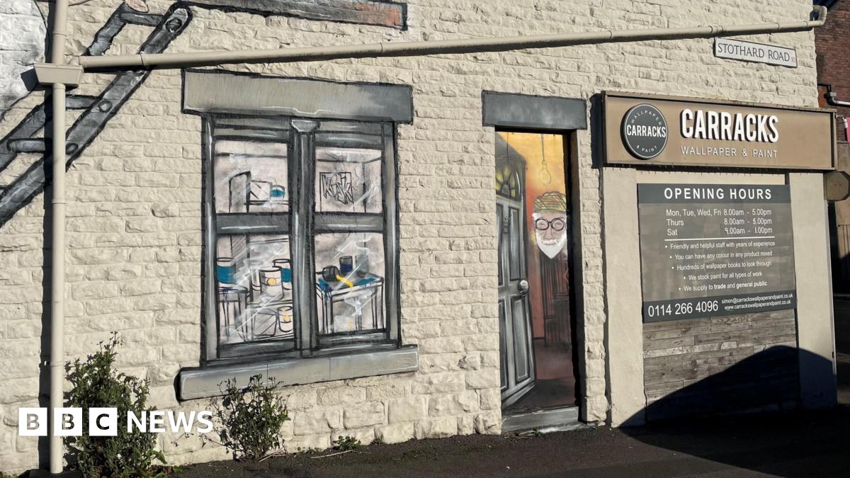 The exterior of bricked building. There is a large mural of a window  painted in a realistic style. To the right of the window mural is a painted door featuring a portrait of an older person wearing glasses and a hat. Above the door is a black and white sign that reads 'Carracks Wallpaper & Paint'.