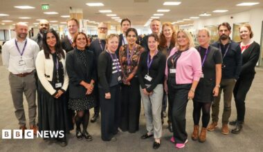 A group of adults gathered in an open conference room. They are smiling and wearing purple lanyards. They are all dressed in office wear.