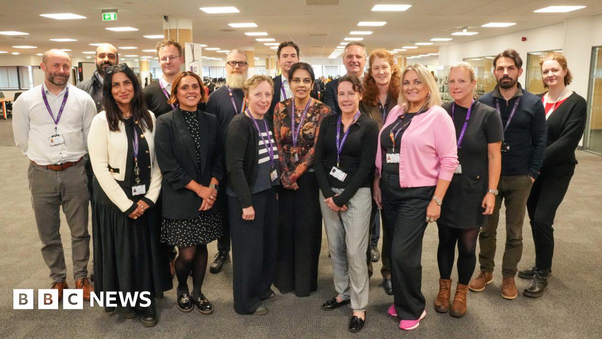 A group of adults gathered in an open conference room. They are smiling and wearing purple lanyards. They are all dressed in office wear.