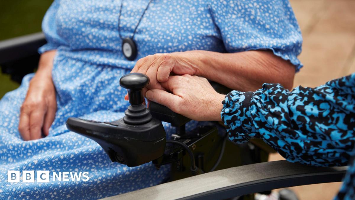 A picture of a women in a wheelchair holding hands with another women. Both are wearing blue patterned dresses and only their hands and part of a chair and a wheelchair can be seen.