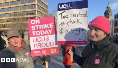 Two men are standing in the city centre. One man in a baseball cap and denim jacket has a placard in pink and white saying Strike Today UCU and Proud. A man next to him has a pink beanie hat and green coat and is holding a placard saying Too Burnout To Think Of A Slogan