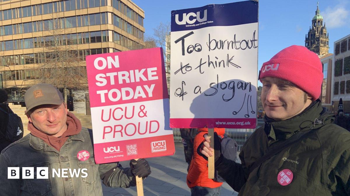 Two men are standing in the city centre. One man in a baseball cap and denim jacket has a placard in pink and white saying Strike Today UCU and Proud. A man next to him has a pink beanie hat and green coat and is holding a placard saying Too Burnout To Think Of A Slogan