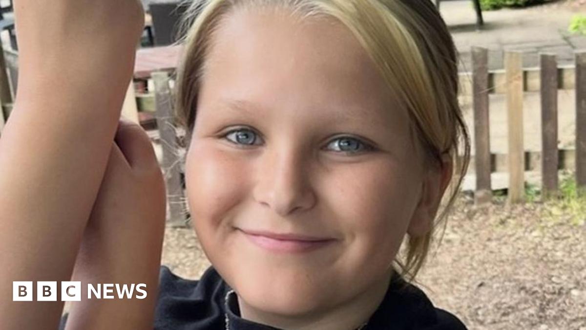 A smiling girl with blue eyes and fair hair pulled back into a ponytail. She is wearing a black top. She is outdoors and there is a wooden fence behind her.