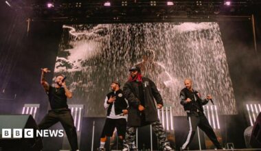 The four members of JLS on stage singing. There is a screen behind them playing a music video with rain pouring down. They are in black clothes and have handheld black microphones