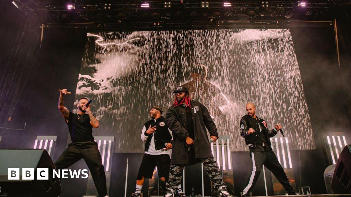 The four members of JLS on stage singing. There is a screen behind them playing a music video with rain pouring down. They are in black clothes and have handheld black microphones