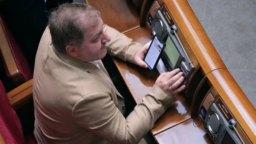 An aerial shot of a brown haired man wearing a cream suit who is sat at a desk looking in the distance while holding a mobile phone.