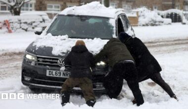 heavy snow on the ground and road with three people helping to push a car that has clearly got stuck