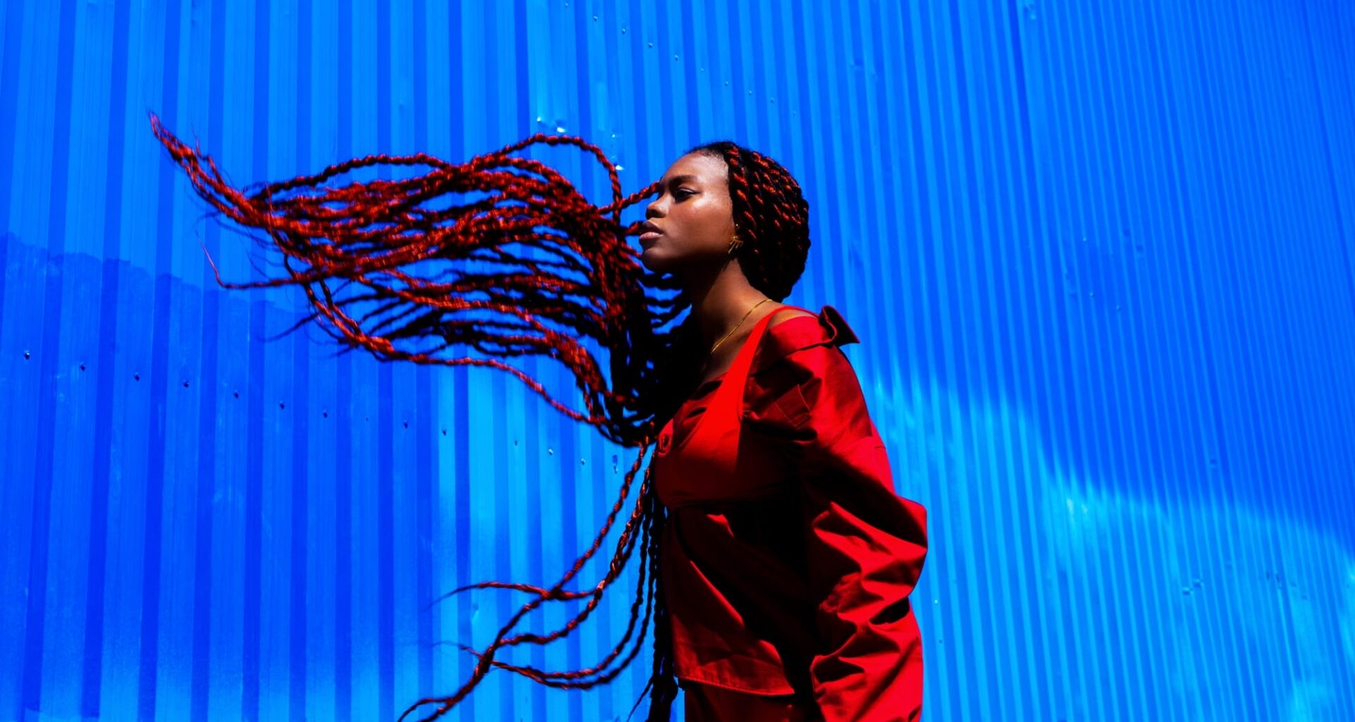a photograph by Yannis Guibinga of a Black woman with long black-and-red braids, wearing a red outfit and standing in front of a bright blue wall