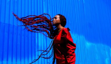 a photograph by Yannis Guibinga of a Black woman with long black-and-red braids, wearing a red outfit and standing in front of a bright blue wall