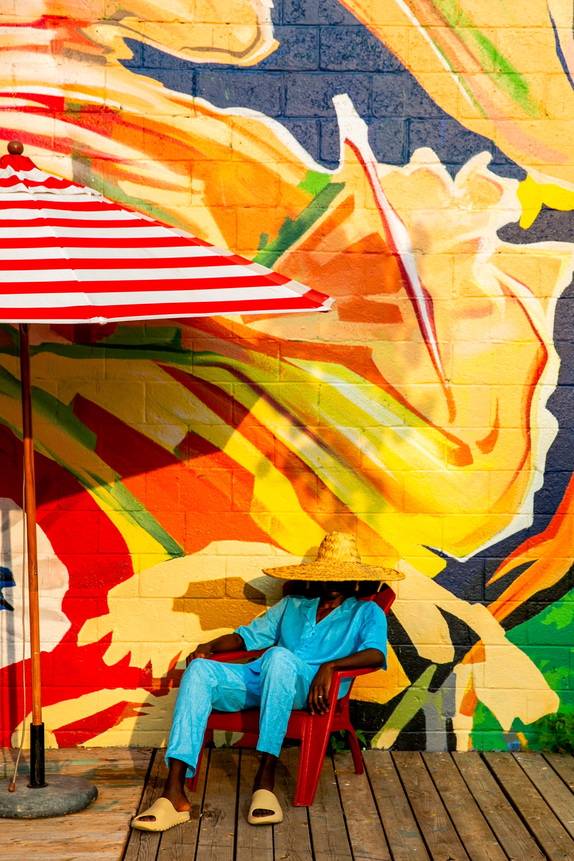 a photograph by Yannis Guibinga of a figure lounging in front of a colorful mural, next to a red-and-white striped umbrella