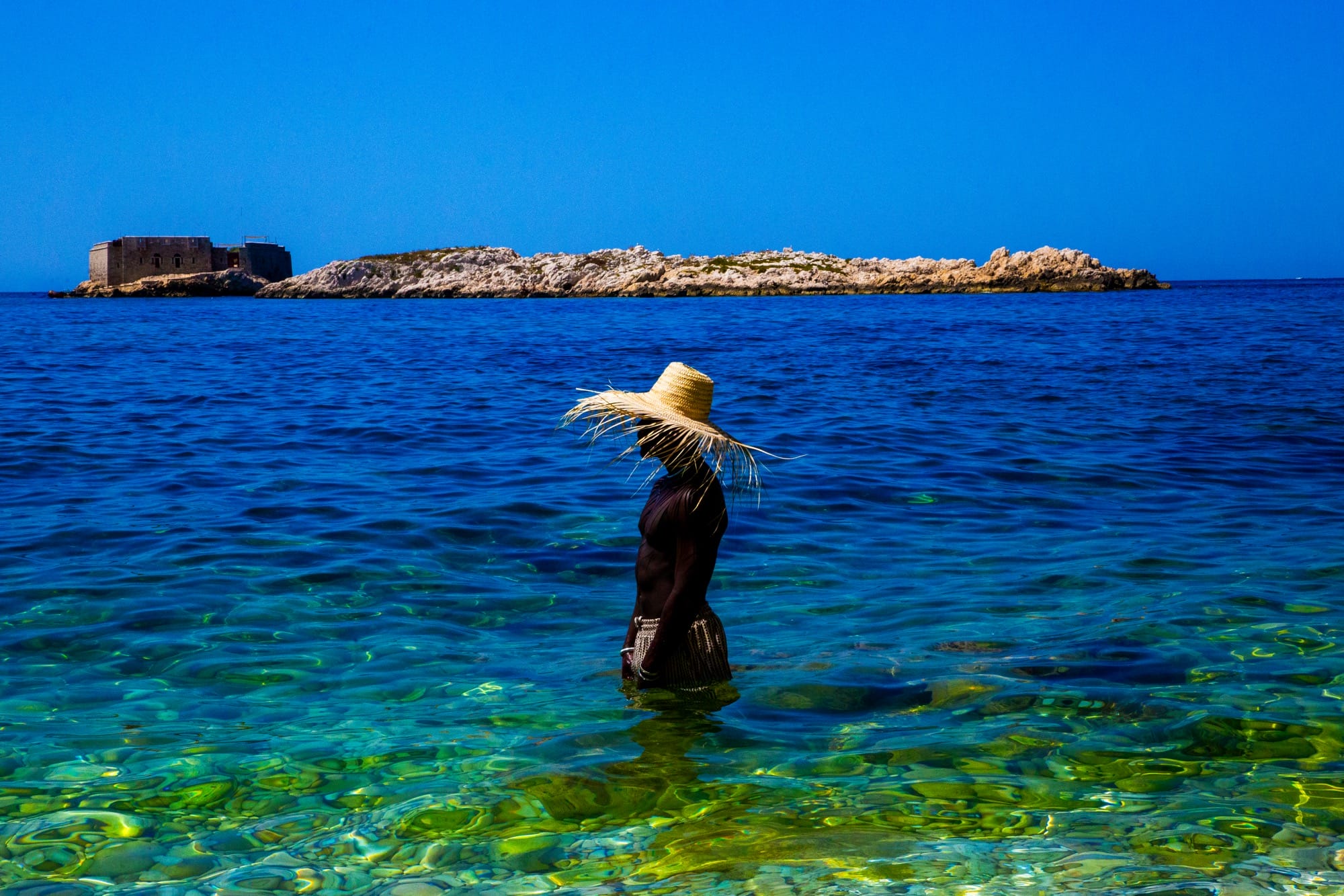 a photograph by Yannis Guibinga of a figure wearing a wide-brimmed hat, standing in the water against a bright blue sky
