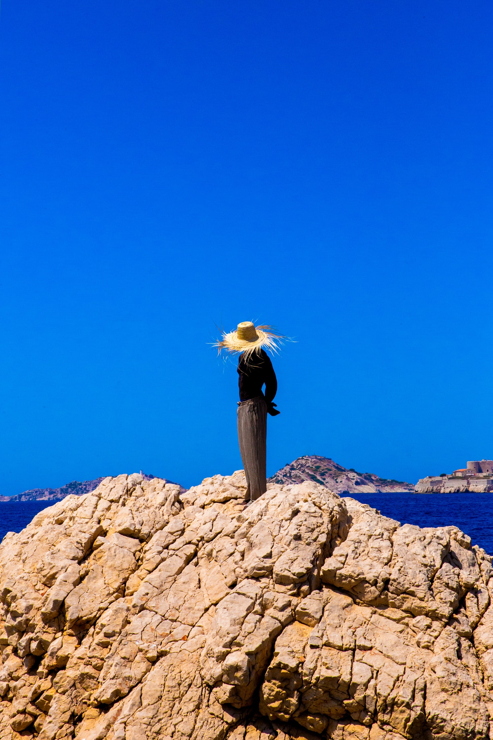 a photograph by Yannis Guibinga of a figure wearing a wide-brimmed hat, standing on a rock in front of a bright blue sky