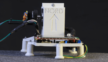 A 3D-printed assembly standing on short legs is visible. A portion extends upward with the word "Nord" sunk into it. Cables extend from one side of the upright portion, and a side view of a circuit board is visible at the front of the assembly.