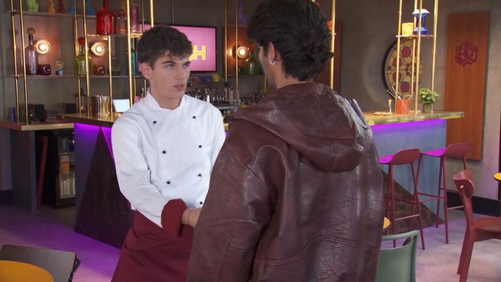 hollyoaks lucas hay and dillon ray in the hutch, lucas wearing his chef white hands a phone over to dillon in a leather jacket