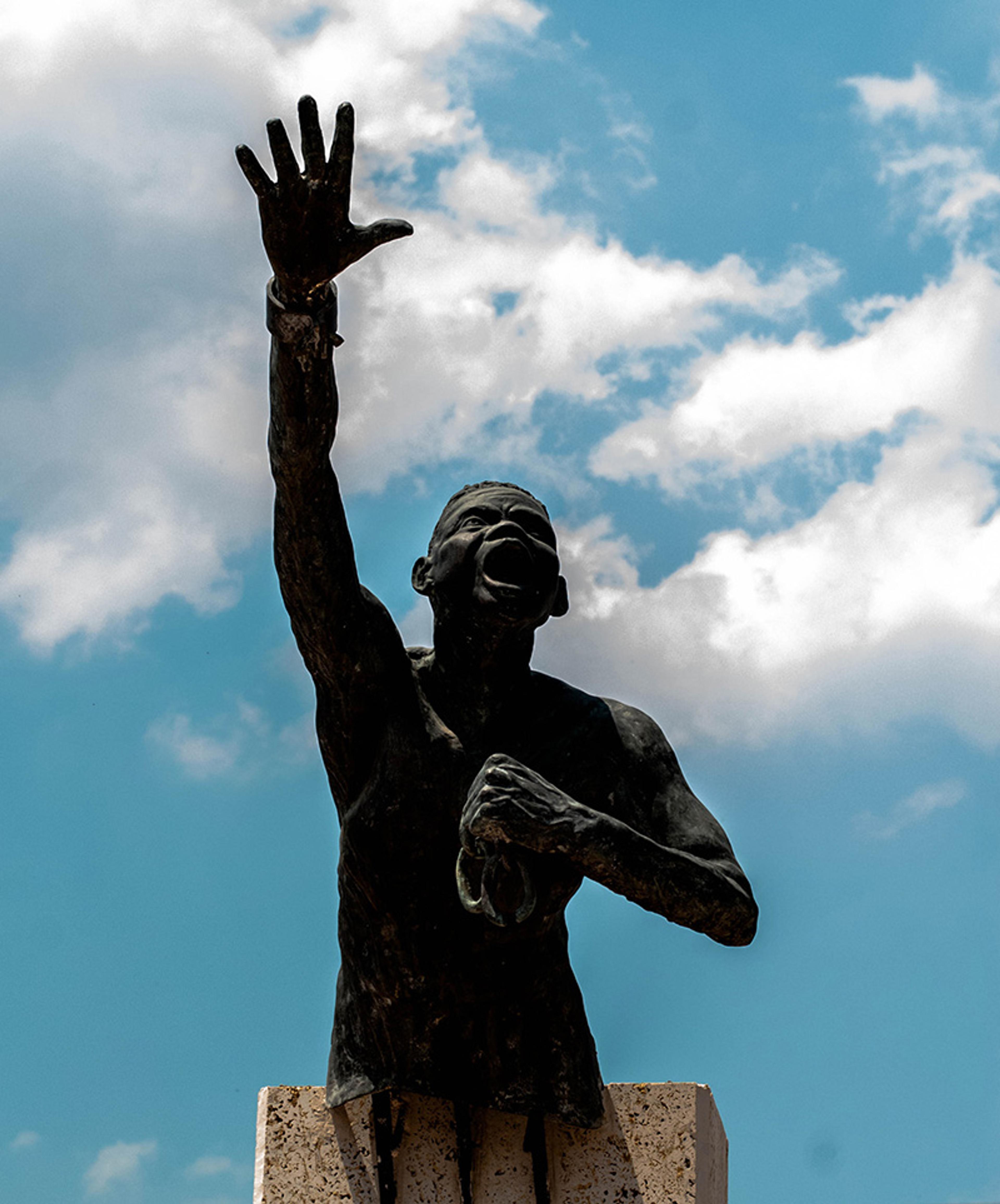 Photo of a statue depicting a person with an arm raised against a backdrop of blue sky and clouds.