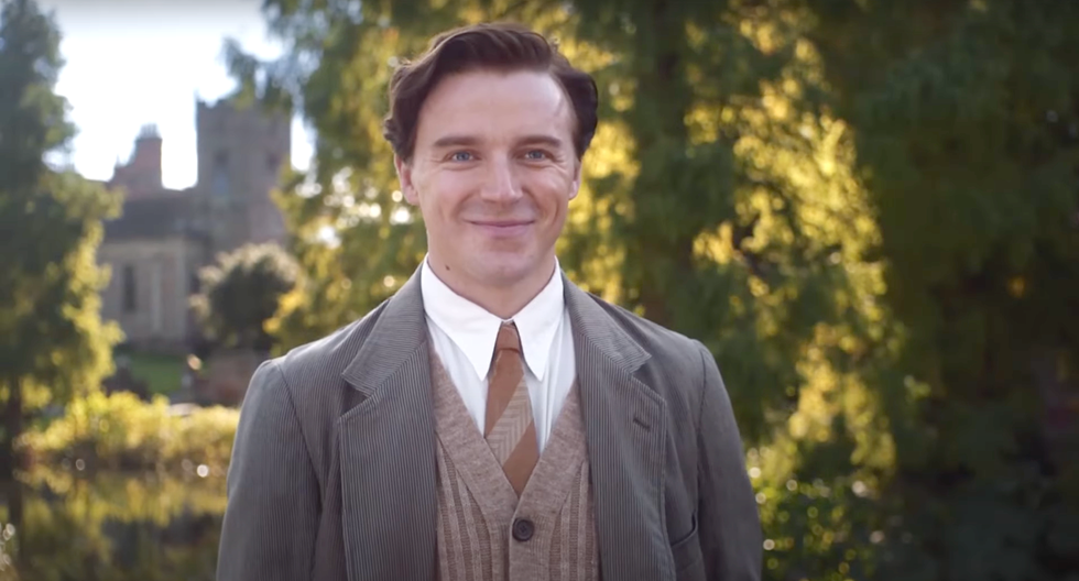 Jack Lowden, Benediction jack lowden walking in a wooded garden on a large estate in benediction, a young man with short hair, smiling, wearing a white shirt, brown jumper and tie with grey coat