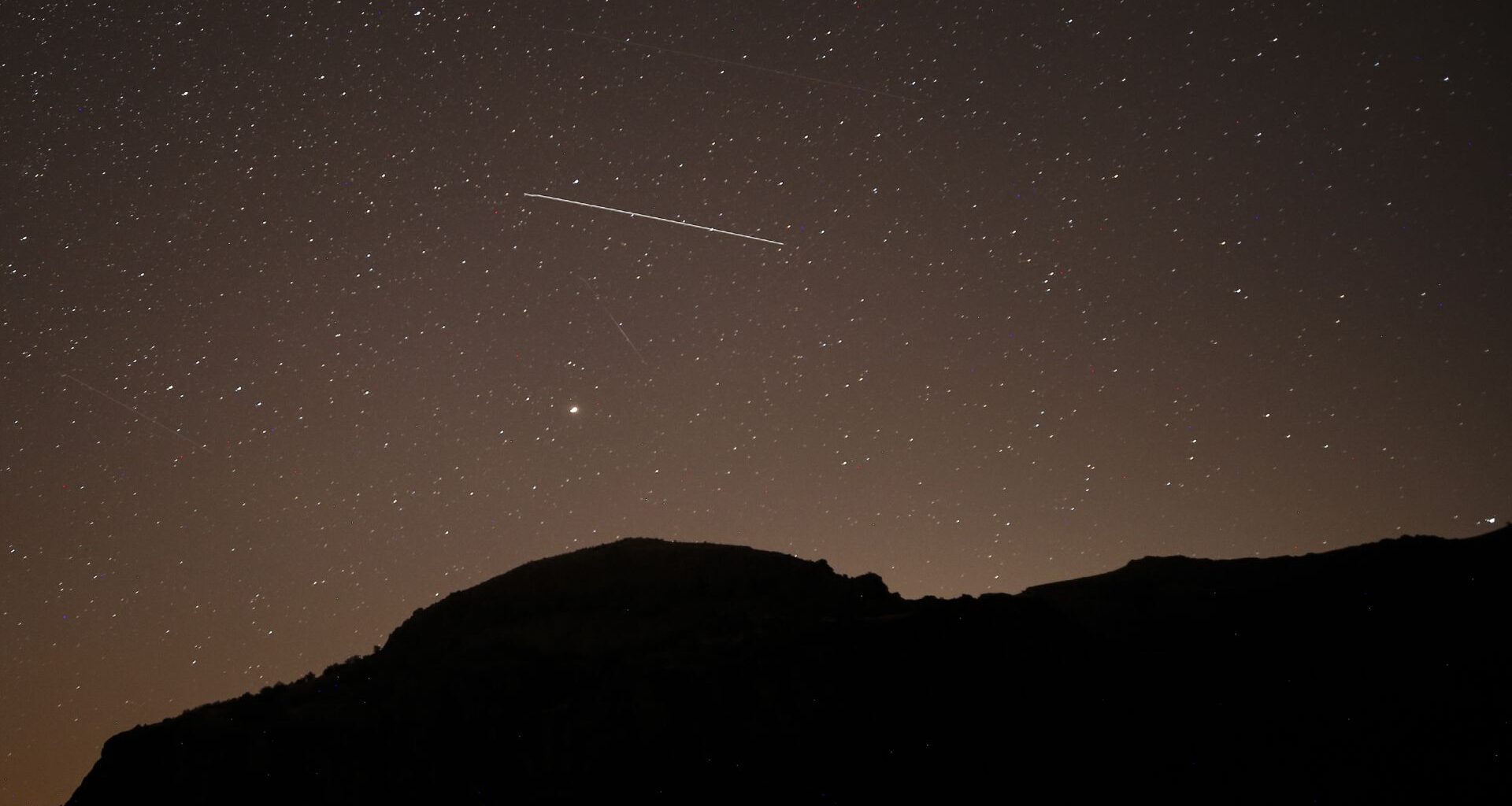 A lone meteor is spotted streaking through a dark starry sky above a silhouetted hill.