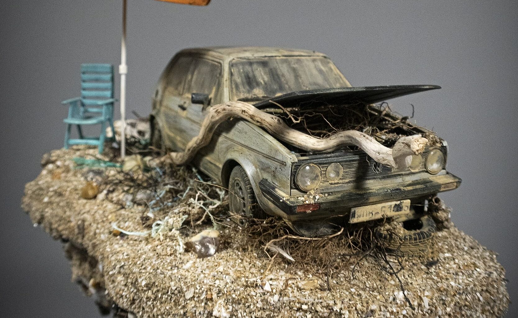 a hand-sculpted miniature by Simon Laveuve of an abandoned car with a large root growing through the hood, with a beach umbrella propped up next to it