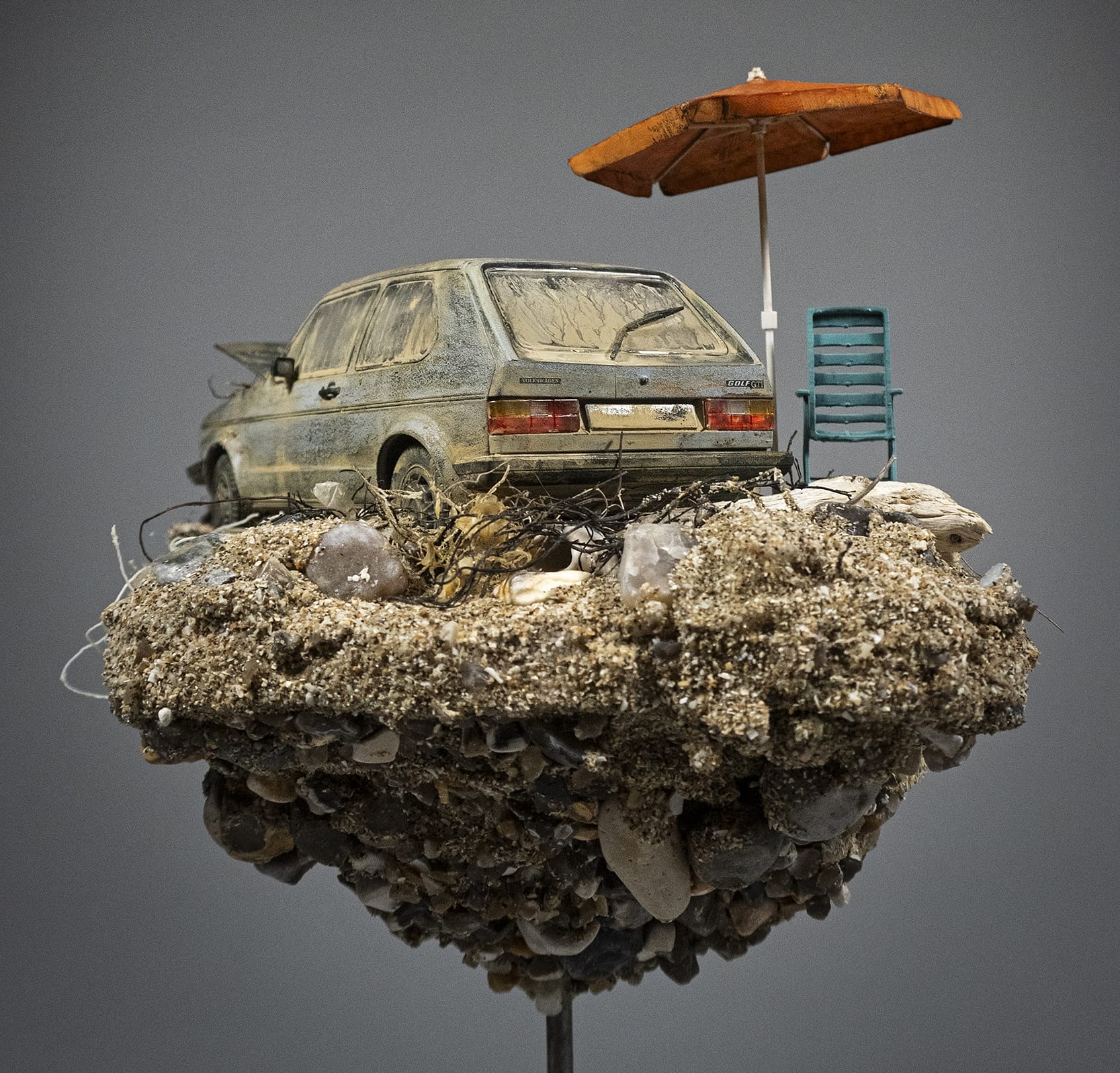 the rear view of a hand-sculpted miniature by Simon Laveuve of an abandoned car with a large root growing through the hood, with a beach umbrella propped up next to it