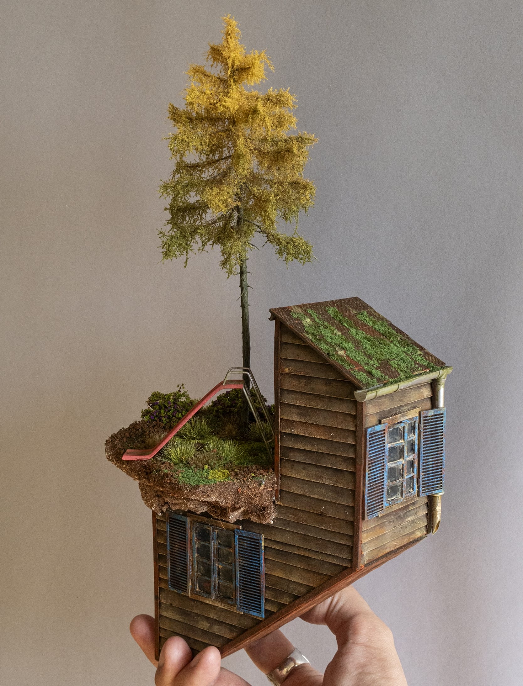 a hand-sculpted miniature by Simon Laveuve, held by the artist's hand for scale, of a shack-like building with a tree and a children's slide