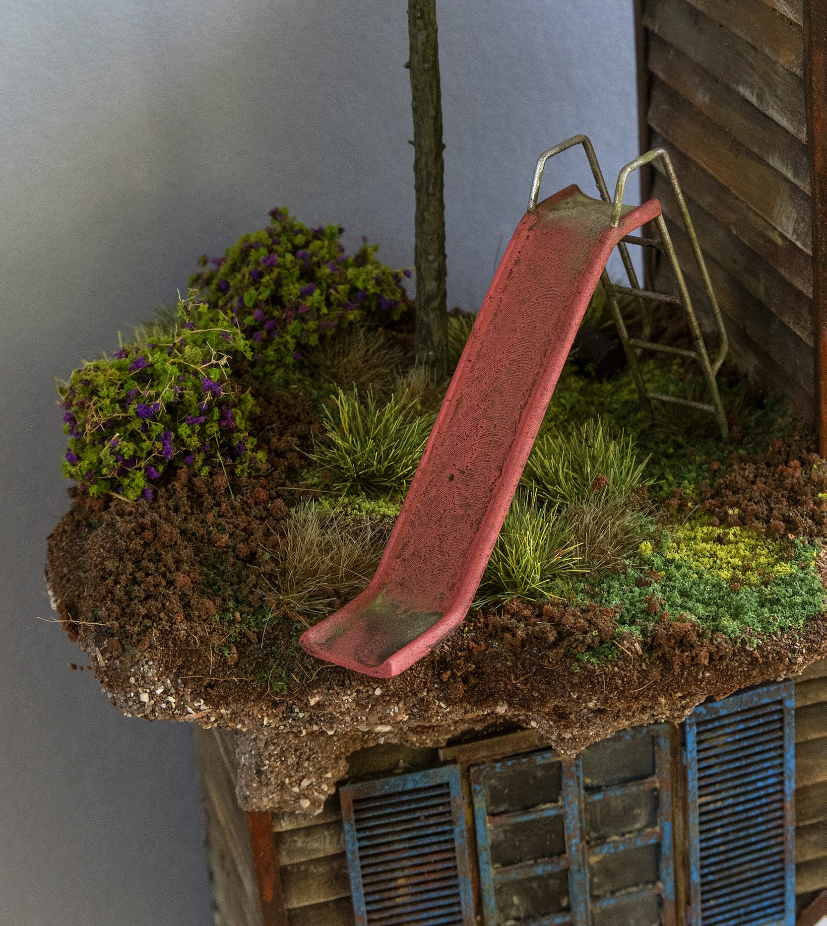 a detail of a hand-sculpted miniature by Simon Laveuve focused on a children's slide nestled in a mossy yard