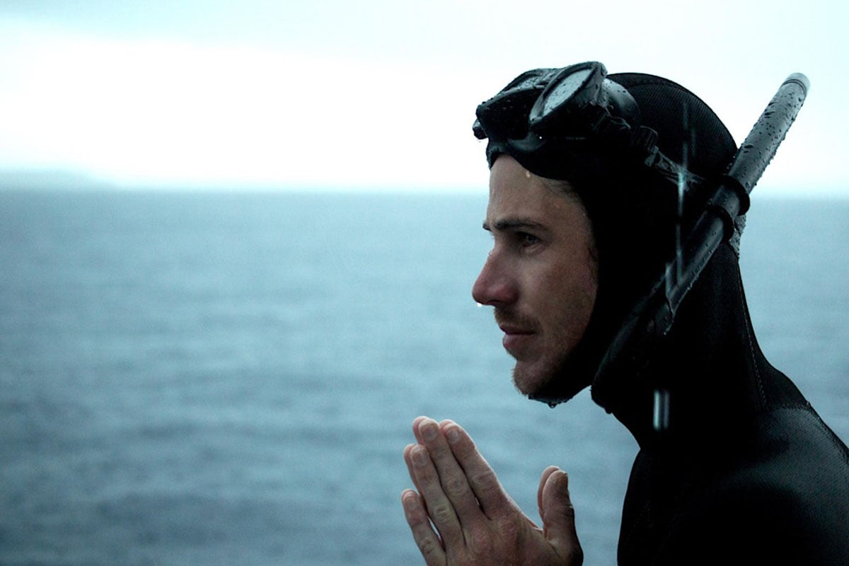 A person in a black wetsuit and snorkel mask stands near the ocean with hands pressed together, appearing focused and contemplative, with the sea and cloudy sky in the background.