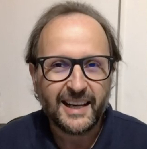 A balding man with a beard wearing dark glasses and a navy shirt looks at the camera