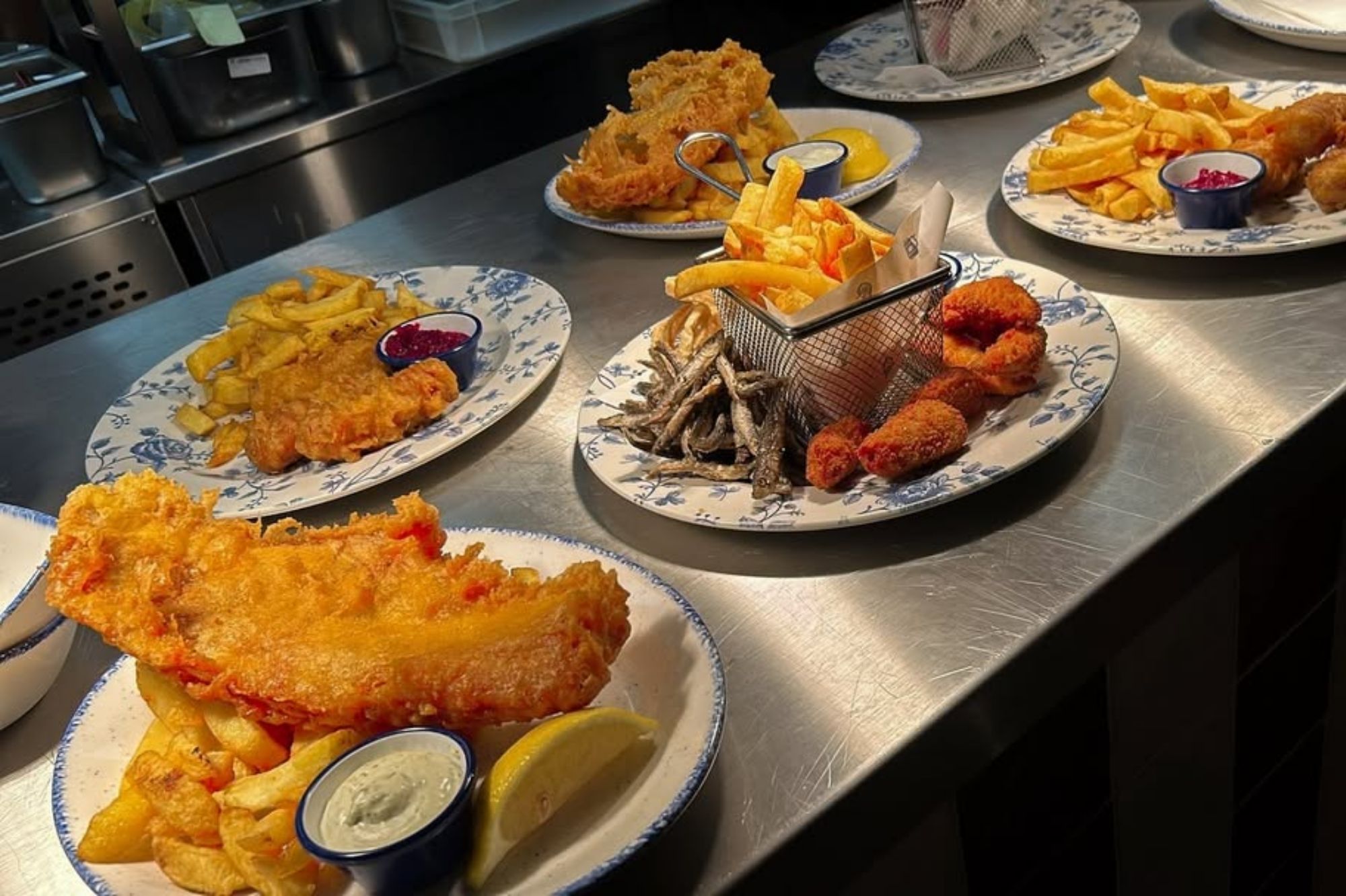 An image collage containing 1 images, Image 1 shows Various plates of fish and chips, battered sausage, and other fried seafood on a metal counter