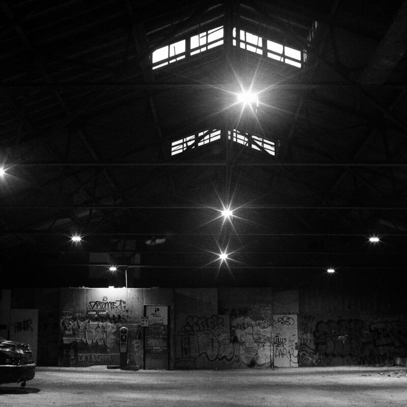 A black and white photo of an empty, dimly lit warehouse with graffiti-covered walls, bright ceiling lights, and windows near the roof. A parked car is partially visible on the left side.
