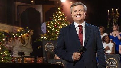 A man holding a microphone stands in a grand hall, with festive trees and lighting in the backdrop. Choir singers can be seen just behind him with candles and Songs of Praise logos embossed crossed the furniture.