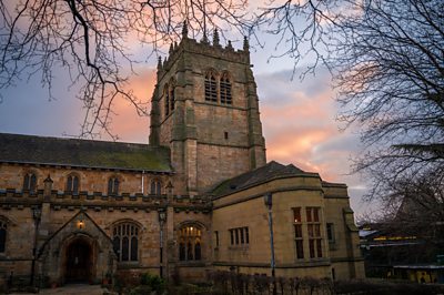 A grand brown cathedral (Bradford Cathedral) stands proudly as pink clouds float behind it. The image is framed with bare winter trees.