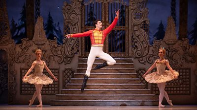 A male ballet dancer leaps high above an ornate stage, dressed as a soldier in a red coat with gold brocade. Behind him, two female ballet dancers strike a pose in gowns with wide skirts.