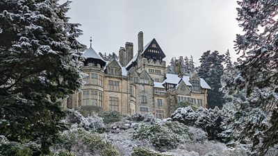 A large house - National Trust Cragside - is visible high on a hill, surrounded by snow dusted trees.