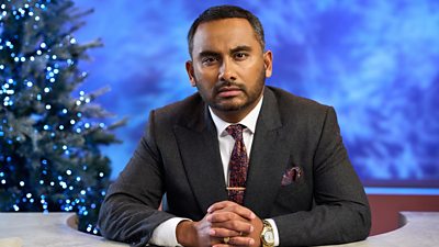 A man in a dark suit, white shirt and dark patterned tie rests his hands on a desk looking series. A Christmas tree with twinkling lights is visible behind him.