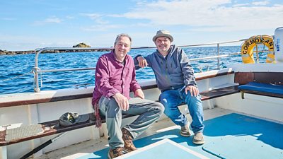 Two men sit together smiling on a bench on a boat at sea on a bright sunny day