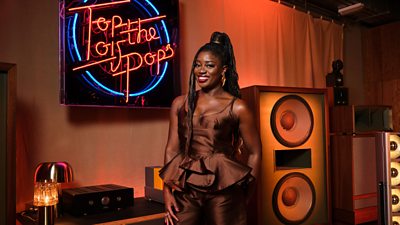 A woman in a brown peplum top and trousers smiles, posing against a giant speaker and a neon Top of The Pops sign