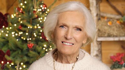 A woman in a light cream jumper smiles to camera, with twinkling Christmas lights on a tree visible behind her