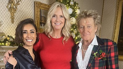Two women and a man pose together smiling beside a fireplace