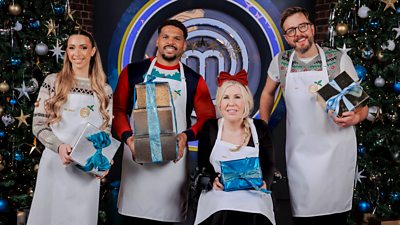 Four celebrities sit and stand in the MasterChef kitchen, all wearing white aprons. They pose between two Christmas trees against a large M MasterChef logo holding presents.
