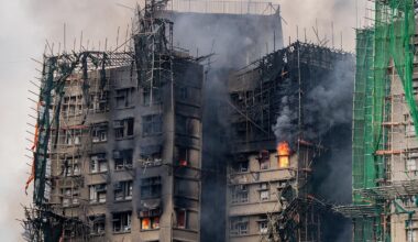 A photograph of one of the high-rise buildings in the Wang Fuk Court complex engulfed in flames and thick black smoke. The structure is covered in bamboo scaffolding, some of which is partially collapsed or charred. Bright orange flames are visible through several windows and along the exterior, while green safety netting hangs loosely on parts of the scaffolding. The sky is filled with dense smoke, obscuring the background.