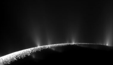 A black and white image of Enceladus' south pole. Several plumes of water ice spray up from the curved surface; they are visible as misty white spikes. Most of the south pole is dark, but a small sliver is lit up at bottom left.