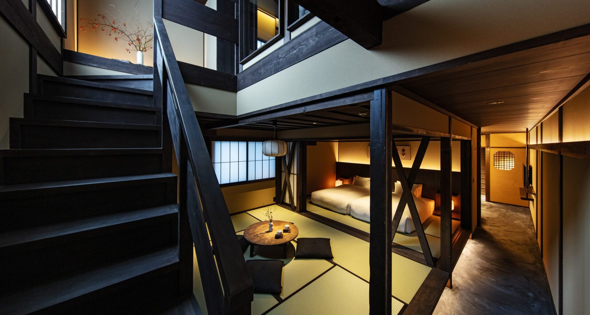 A wooden staircase and banister go up the left side of the photo, with a vase and flowers at the top of the stairs. A warmly lit, open plan area contains a small coffee table with futon-cushions for sitting and two beds towards the back of the space.