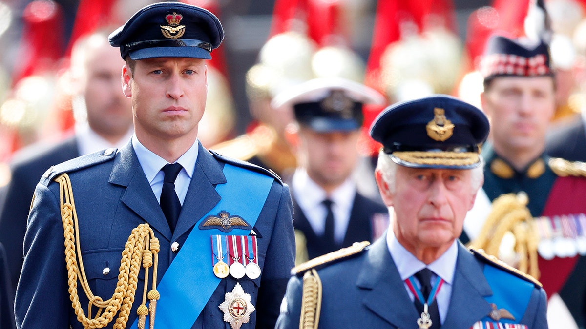 Prince William in a military suit walking behind his father King Charles also in a military suit.