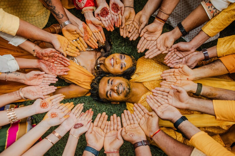 Two people in yellow outfits lie on grass, smiling, surrounded by a circle of friends showing open hands decorated with henna designs, bracelets, and colorful clothing, creating a festive, joyful scene.